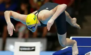 An Australian swimmer in a yellow cap prepares for a powerful high diving start, poised on the edge of the block.