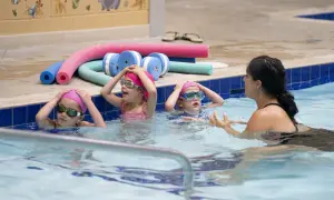Children in a pool during youth swim lessons, wearing pink caps and goggles, with a swim instructor.