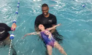 A smiling instructor helps a young girl in a Frozen swimsuit learn how to float on her back in a swimming pool.