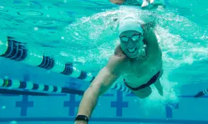 A smiling man swims underwater in a pool, enjoying easy swim workouts. Lane lines and pool markings are visible.