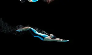 Swimmer in a blue and black suit performs a powerful dolphin kick underwater, creating a trail of bubbles against a dark background.