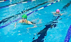 Two swimmers in a pool, one in a green cap doing breaststroke, the other in a pink cap swimming freestyle.