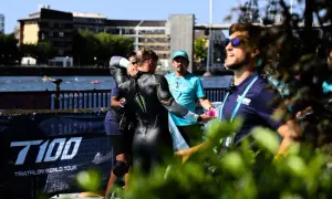 A triathlete in a wetsuit is helped by staff during a triathlong transition. The T100 Triathlon World Tour banner is visible.