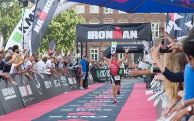 A triumphant athlete crosses the finish line at an Ironman triathlon, arms raised in victory, cheered by a crowd.