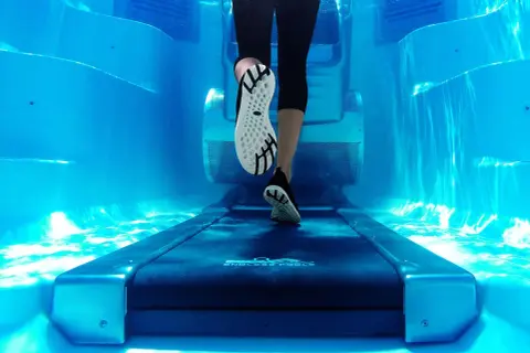 Underwater view of a person's legs and feet on a treadmill, performing aqua jogging in a bright blue pool.