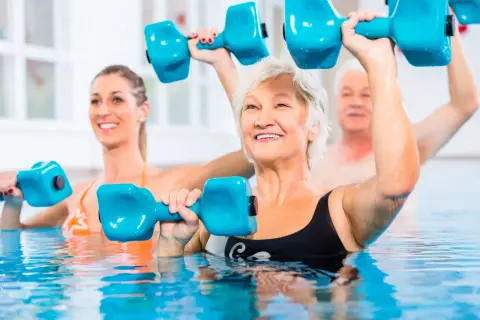 Seniors and a younger woman enjoy pool exercises, lifting blue dumbbells for a refreshing workout.