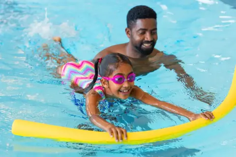 A father smiles as he helps his daughter with her swim lessons. She's wearing goggles and holding a yellow pool noodle.