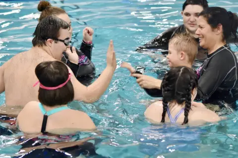 A group enjoys swim lessons for special needs, with instructors and participants high-fiving and smiling in the pool.