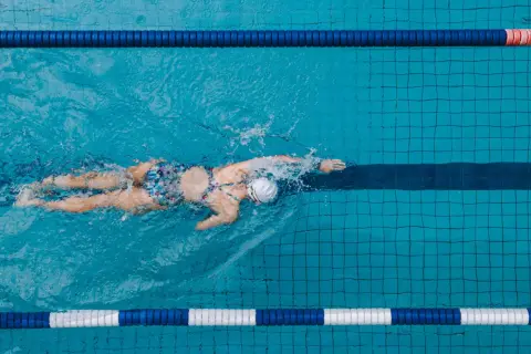 A lone swimmer glides through a pool lane, their back and arms creating splashes. The colorful swimsuit stands out against the blue water.