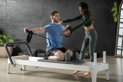 A man on a reformer uses resistance bands for pilates for swimmers, with a trainer guiding his form.