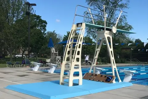 A tall, cream-colored high diving board stands ready for action next to a bright blue pool.