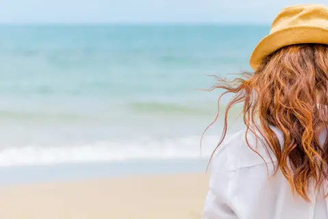 Woman with red hair in a straw hat on the beach, wondering how soon can I swim after coloring my hair.