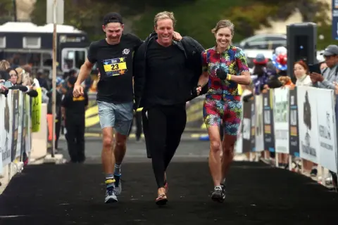 Three friends celebrate crossing the finish line of an Ironman relay. The woman in the colorful floral suit beams, while the two men, one in a black shirt with a race bib, share a laugh.