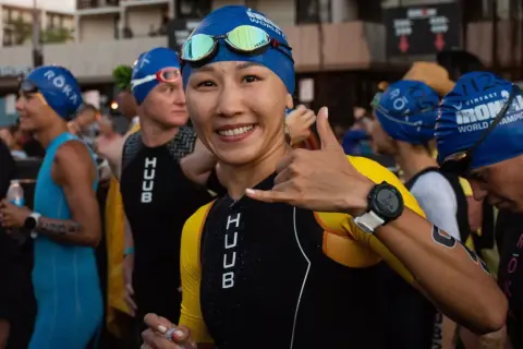 A smiling woman in a blue swim cap and yellow and black triathlon suit gives a shaka sign, ready for the honu triathlon.