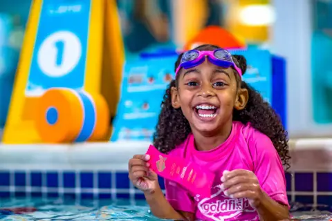 A happy girl in goggles and a pink rash guard holds a Goldfish Swim School ribbon. She's in a pool, ready for swim classes for 5 year olds.