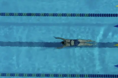 A swimmer glides through the water, executing a powerful breaststroke. The blue pool is marked with lane lines and colorful flags.