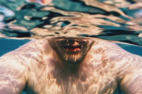 Underwater shot of a man's shoulders and mouth, with light patterns on his skin. A visual guide on how to breathe underwater for beginners.