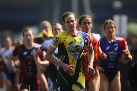 Katie Zaferes, number 01, leads a pack of triathletes in a race, her yellow and black suit featuring a rhino logo.