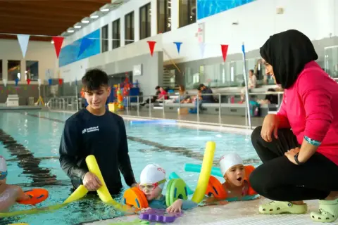 A swim instructor in a pink shirt and hijab coaches young children in a pool, demonstrating the rewarding nature of swim instructor jobs.