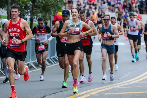 Runners in a marathon, some wearing "Greater Boston" gear, push through the race. The image captures the intensity of the iron man marathon length.