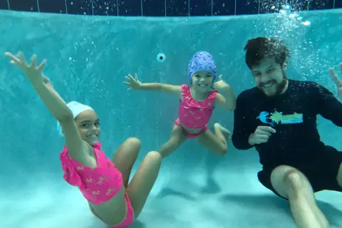 A swim instructor smiles while teaching two young girls how to teach toddler to hold breath underwater.