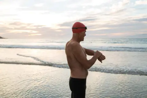 Man in swim cap checks watch on beach, ready for his swim. Early morning or late evening might be the best time to swim for weight loss.