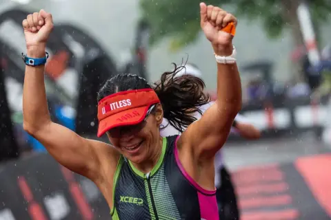 A triumphant athlete celebrates crossing the finish line of an Ironman length race in the rain.