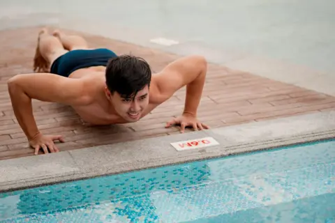 Man doing push-ups on a wooden deck next to a swimming pool.