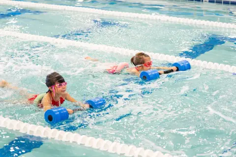 Two girls with goggles swim in a pool, holding blue flotation devices. One girl's face is partially submerged, showing her determination.