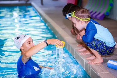 Instructor demonstrates water pouring during swim lessons for teens. Children watch intently from the poolside.