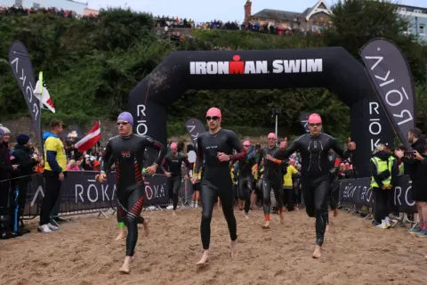 Athletes in wetsuits run from the water under an "Ironman Swim" arch. The start of their race is a sandy beach, ready for the iron man swim challenge.