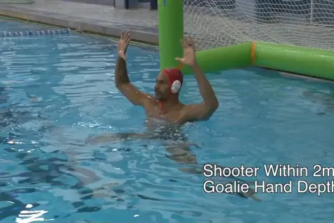 A water polo goalie in a red cap with hands raised, ready to block a shot.