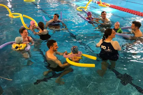 Parents and babies enjoy mommy and me swim classes, splashing and playing with pool noodles in the bright blue water.