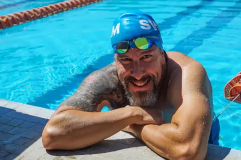 A smiling man in a blue swim cap and goggles rests on the pool edge, ready to teach how to swim laps for beginners.