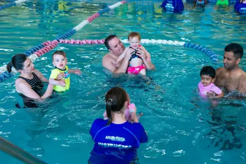 Parents and babies enjoy a swim tots class in a bright blue pool. An instructor in a blue shirt with "Aqua-Tots" visible helps a baby.