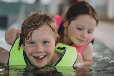 A happy boy with a missing tooth grins during 3 year old swim lessons, with a girl in a pink life vest behind him.
