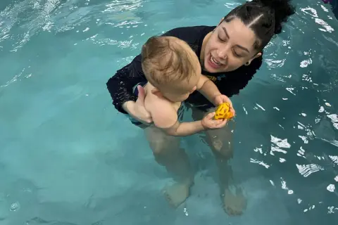 A woman holds a baby in a pool, both smiling. The baby plays with a yellow toy. This scene shows that you can swim with tubes in your ears.
