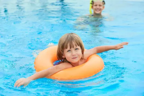 A happy child floats in an orange ring in a pool. Another child in goggles is in the background. This image reminds us when to stop swim lessons: when they're confident and enjoying the water!