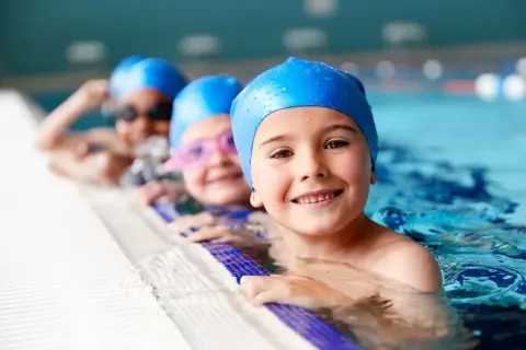Happy kids in blue swim caps at swim lessons for kids, smiling by the pool edge.