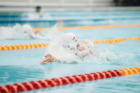 A swimmer in a white cap and goggles powers through the water, creating a splash. Their focused swimmer posture is evident as they propel forward in the lane.