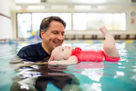 A father smiles as he holds his baby in a pool, showing that kids can learn to swim from a very young age.