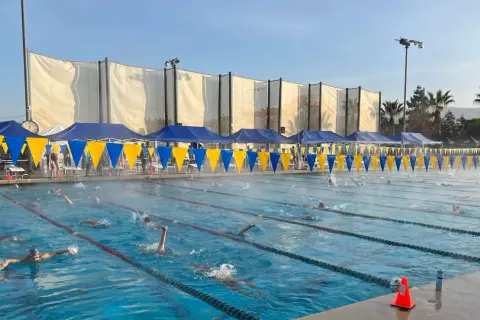 Swimmers race in lanes during a circle swim event. Blue and yellow banners line the pool, with tents and spectators in the background.
