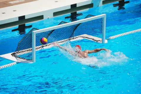 A water polo player in a red cap lunges for the ball near the net, splashing water. Intense underwater games are a thrilling spectacle.