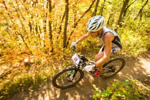 Athlete races through an autumn forest during an off road triathlon, navigating a dirt path with colorful foliage.