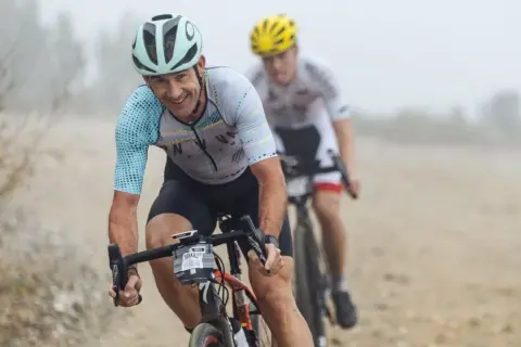 Smiles and determination on the faces of cyclists during a gravel triathlon. The lead rider, wearing a light blue and white jersey, grins as he navigates the dusty course.
