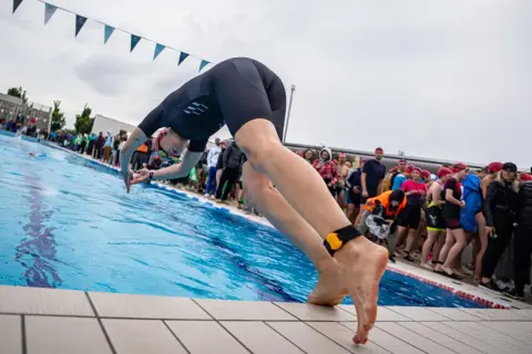 A swimmer in a black wetsuit dives into a pool, ready for advanced swim workouts. Spectators watch from the side.