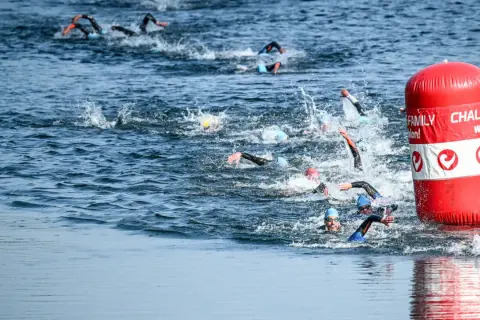 Athletes compete in a triathlon, their triathlon swim strokes creating splashes as they navigate the water around a red buoy.