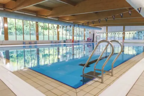 Indoor swimming pool with large windows overlooking trees. Considering private swim lessons cost? This serene setting offers a peaceful learning environment.