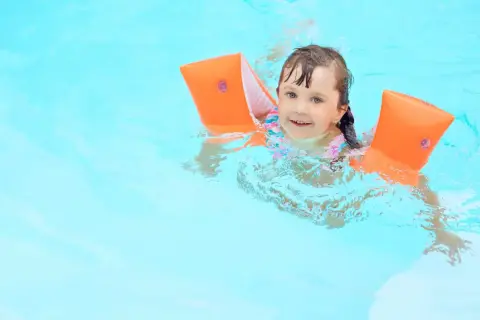 A happy young girl with bright blue eyes is floating in the pool, wearing orange arm floaties and a colorful swimsuit.