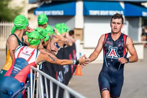 A male NCAA triathlon athlete in a dark blue and red suit with an "A" logo high-fives a competitor.